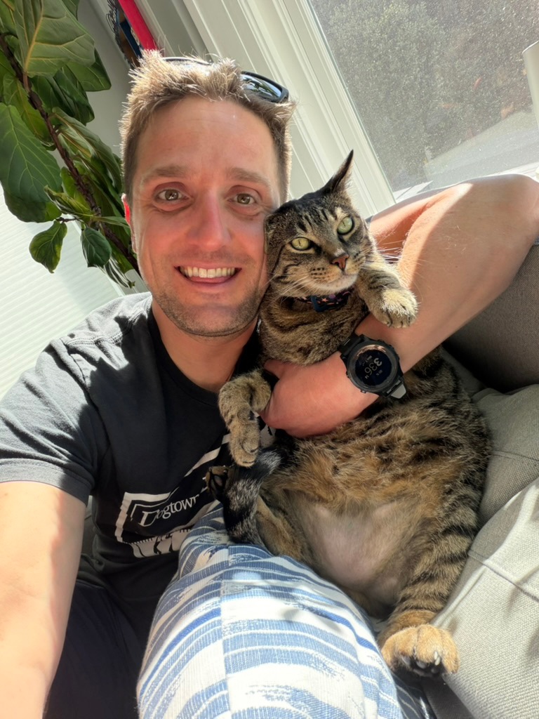 James smiling and holding Baxter, his tabby cat, by a sunny window